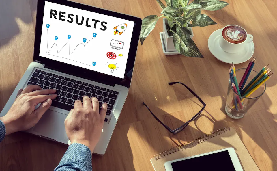 PCを操作する男性の手。めがね、コーヒー・筆記具・ノート。PC画面にはRESULTの文字。