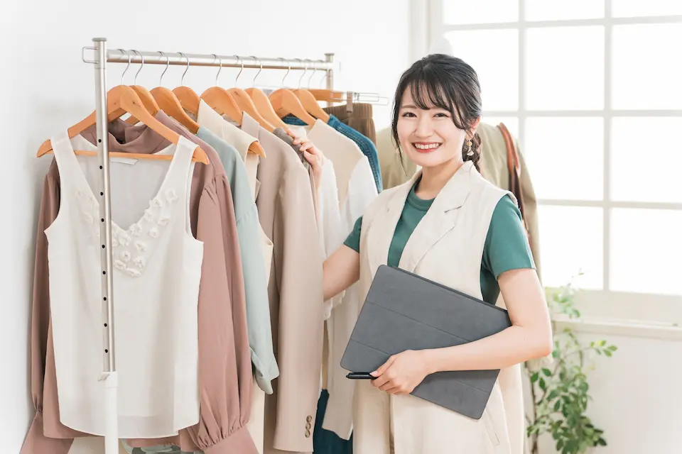 ショップ内でハンガーにかかった衣服に手をかけほほ笑む女性オーナー